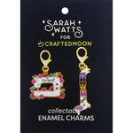 Sarah Watts for Crafted Moon collectable enamel charms on a black display card. The set includes a sewing machine charm decorated with flowers and the word 'therapist', and a colorful geometric animal charm shaped like a bird or fox.
