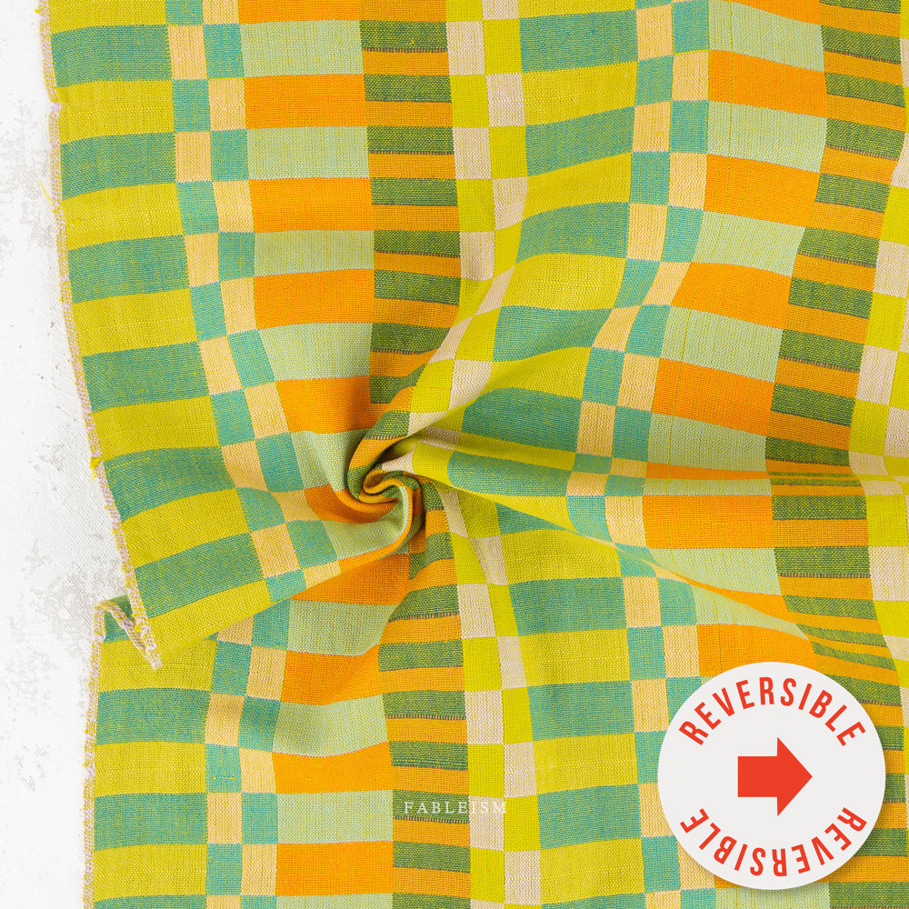 A close-up of a woven fabric with a retro checkered pattern in yellow, orange, green, and beige, featuring a central swirl fold and a white circular sticker overlay that reads 'REVERSIBLE'.