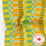 A close-up of a woven fabric with a retro checkered pattern in yellow, orange, green, and beige, featuring a central swirl fold and a white circular sticker overlay that reads 'REVERSIBLE'.