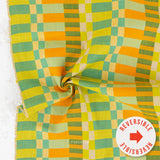 A close-up of a woven fabric with a retro checkered pattern in yellow, orange, green, and beige, featuring a central swirl fold and a white circular sticker overlay that reads 'REVERSIBLE'.