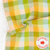 A folded piece of reversible plaid fabric with a soft linen texture, featuring a checkered pattern of green, yellow, peach, and light blue. A white circular sticker in the bottom right corner reads 'REVERSIBLE' with a red arrow.