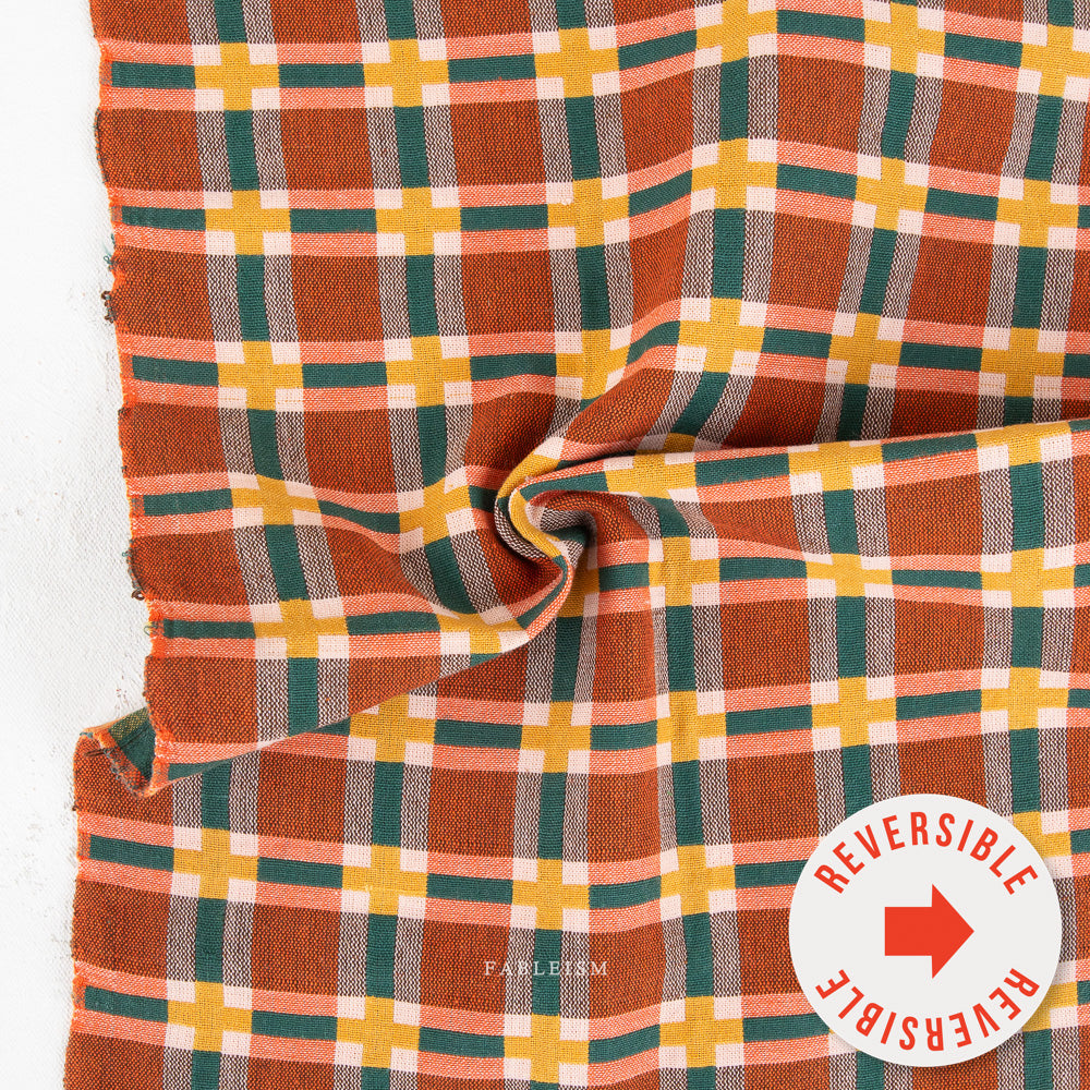 A close-up of a plaid fabric with a warm rust-orange base, overlaid with green, yellow, peach, and white stripes. The fabric is draped in a swirl, showing its texture and pattern.