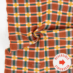 A close-up of a plaid fabric with a warm rust-orange base, overlaid with green, yellow, peach, and white stripes. The fabric is draped in a swirl, showing its texture and pattern.