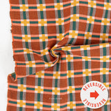 A close-up of a plaid fabric with a warm rust-orange base, overlaid with green, yellow, peach, and white stripes. The fabric is draped in a swirl, showing its texture and pattern.