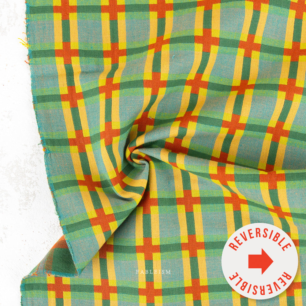A close-up of a plaid fabric with a green, yellow, and orange pattern, draped to show its texture and reversible side, with a white circular sticker overlay in the bottom right corner.