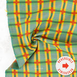 A close-up of a plaid fabric with a green, yellow, and orange pattern, draped to show its texture and reversible side, with a white circular sticker overlay in the bottom right corner.
