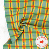 A close-up of a plaid fabric with a green, yellow, and orange pattern, draped to show its texture and reversible side, with a white circular sticker overlay in the bottom right corner.