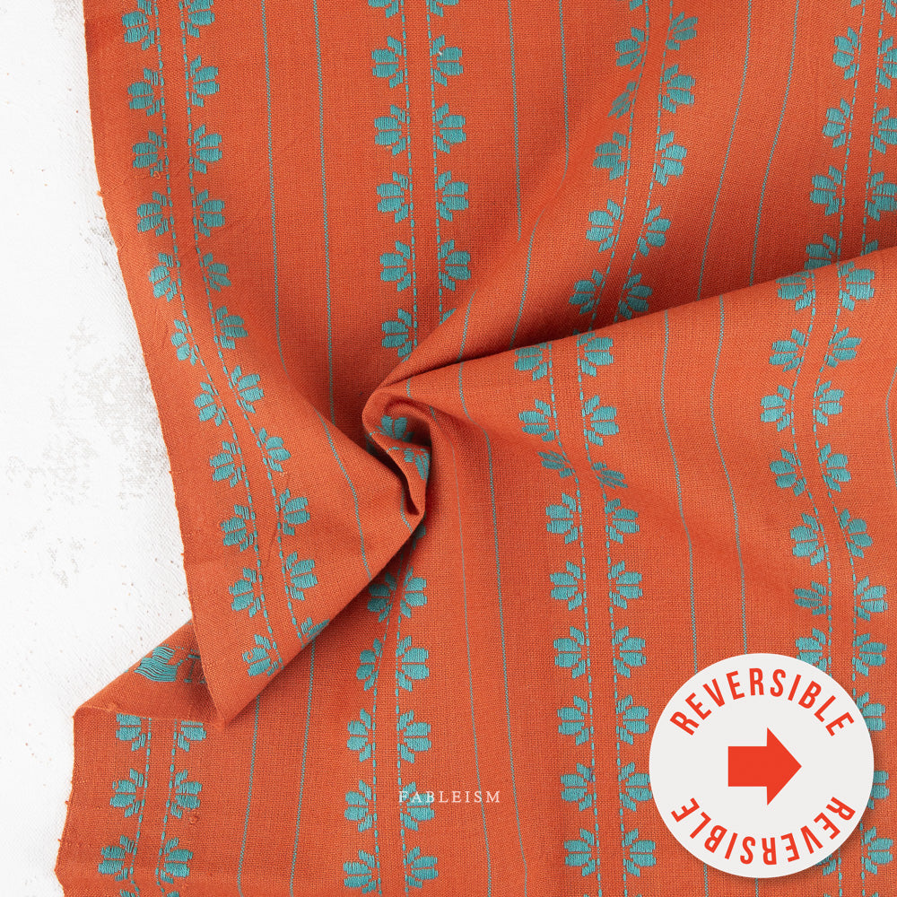 Orange fabric with vertical teal floral motifs and thin teal stripes, draped to show its reversible side, with a white circular 'REVERSIBLE' badge in the lower right corner.