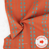 Orange fabric with vertical teal floral motifs and thin teal stripes, draped to show its reversible side, with a white circular 'REVERSIBLE' badge in the lower right corner.