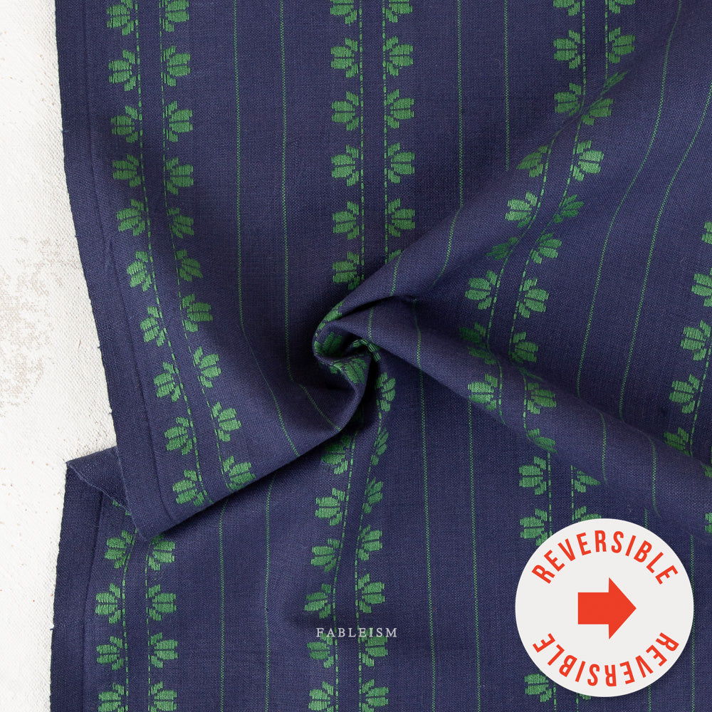 A close-up of a navy blue fabric with vertical green stripes and repeating green floral motifs, draped to show its texture and pattern. A white circular sticker with a red arrow reads 'REVERSIBLE' is placed in the bottom right corner, and the brand name 'RABLBISM' is printed near the bottom center.