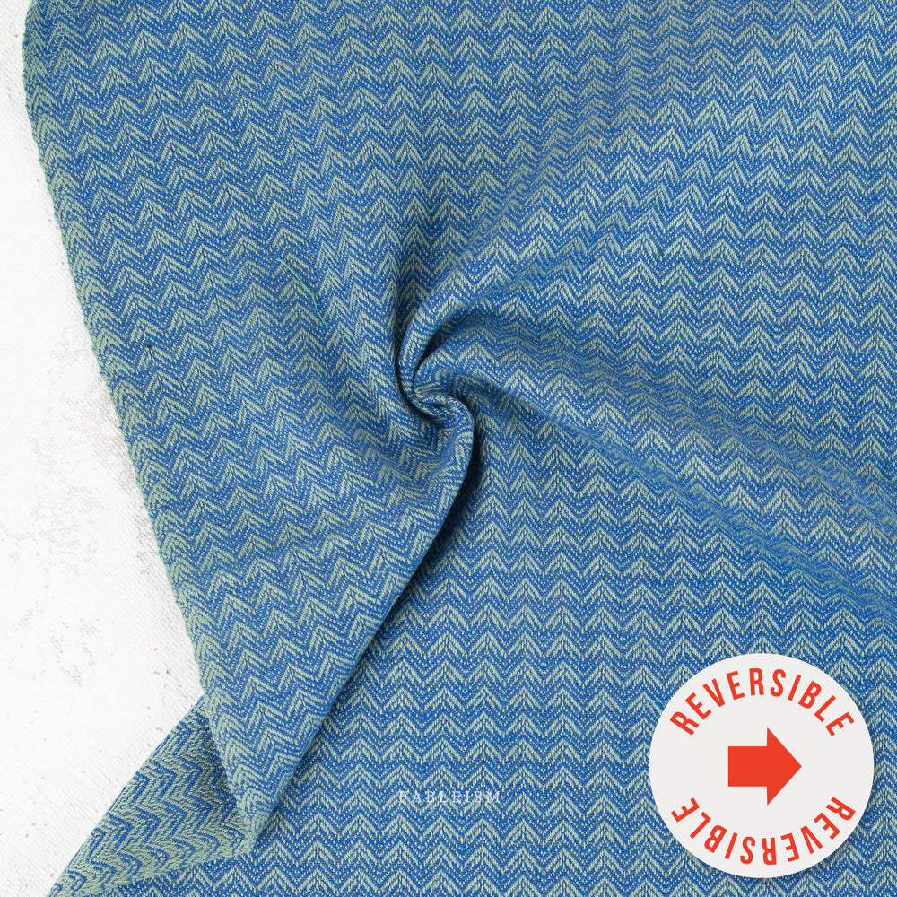A close-up of a blue woven fabric with a repeating chevron pattern, draped to show its texture and reversible side, with a circular 'REVERSIBLE' badge in the bottom right corner.