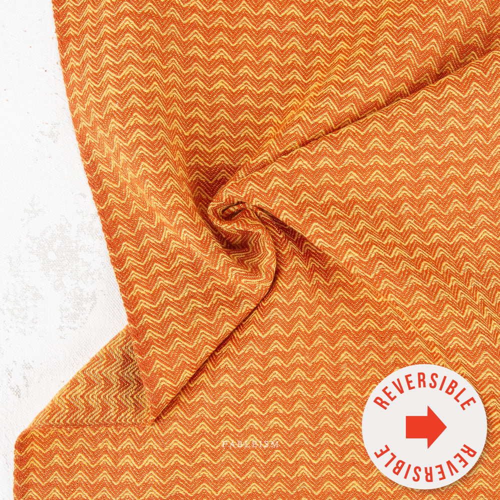 A close-up of folded fabric with a repeating chevron pattern in orange and cream, with a white circular badge in the lower right that reads 'REVERSIBLE' with a red arrow.