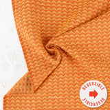 A close-up of folded fabric with a repeating chevron pattern in orange and cream, with a white circular badge in the lower right that reads 'REVERSIBLE' with a red arrow.