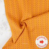 A close-up of folded orange fabric with a repeating small geometric pattern, shown on a light textured surface. A white circular badge in the lower right says 'REVERSIBLE' with a red arrow, and a small 'PABLEISM' mark appears near the bottom center.