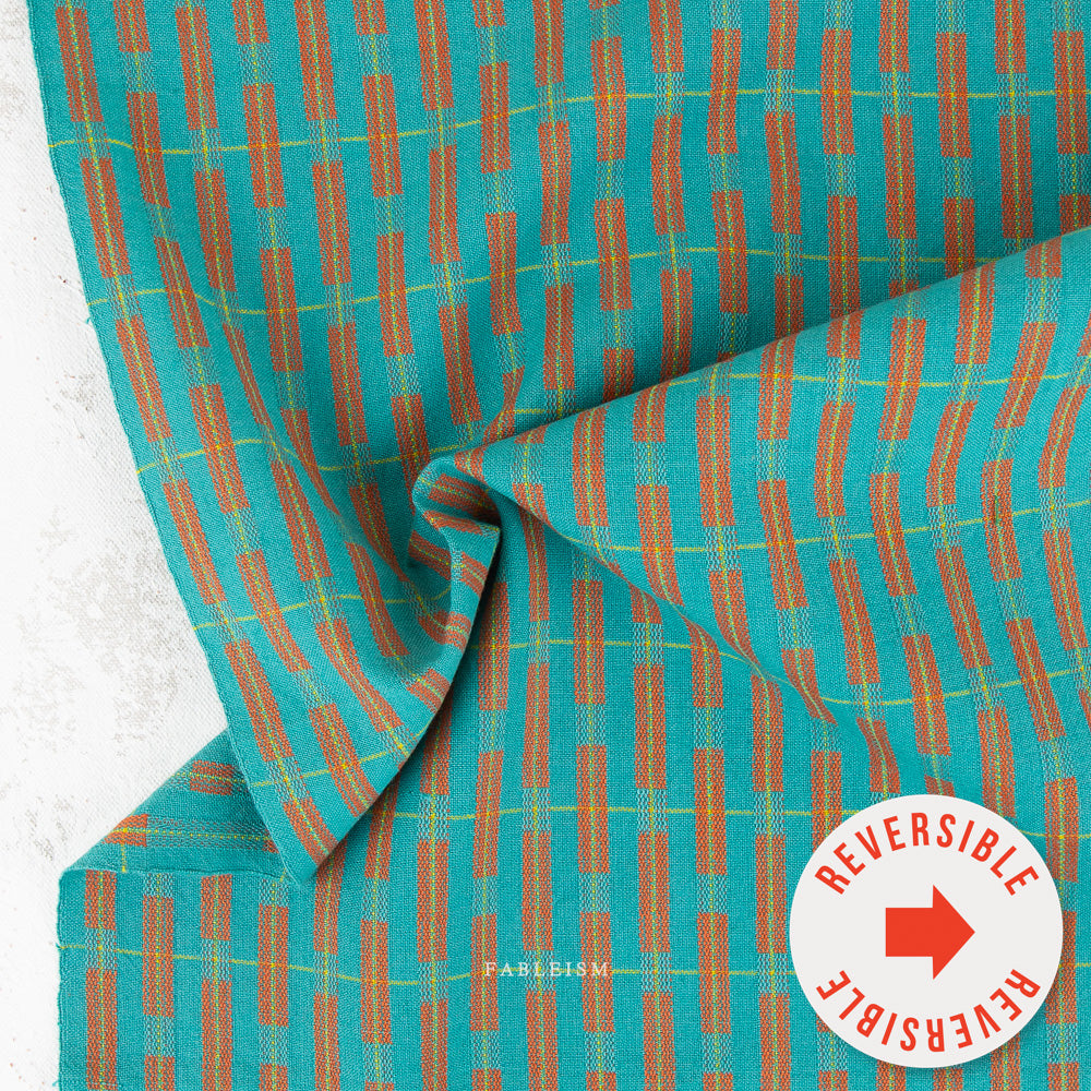 A close-up of a woven textile with a turquoise base and a repeating pattern of rust-orange vertical bars outlined with thin yellow lines. The fabric is draped to show its texture and weave, with a white circular badge in the bottom right corner reading 'REVERSIBLE' with a red arrow.
